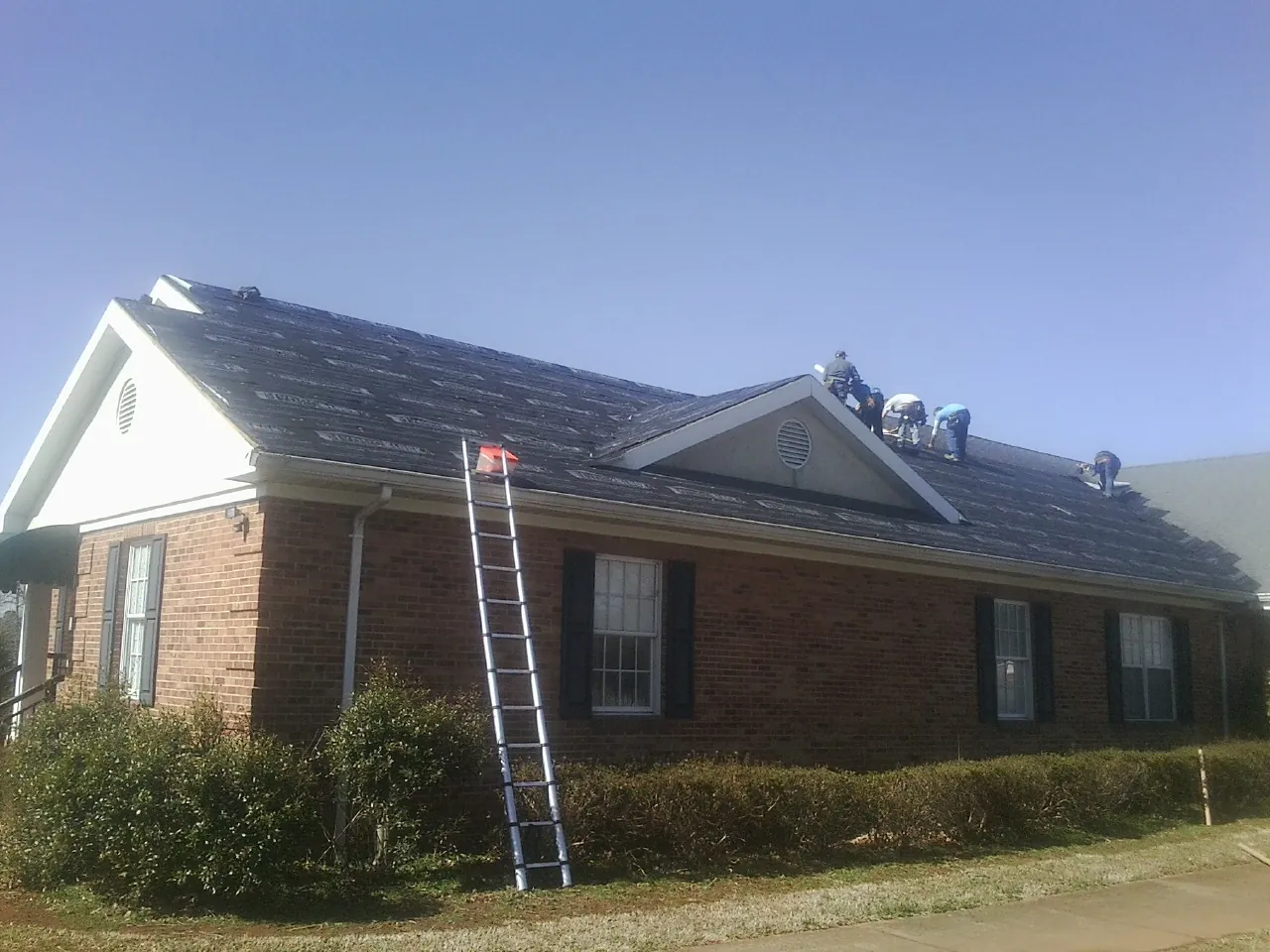 Crew tearing shingles off a brick ranch roof with a ladder set — Emergency Roof Repair work by our crew in Rebecca