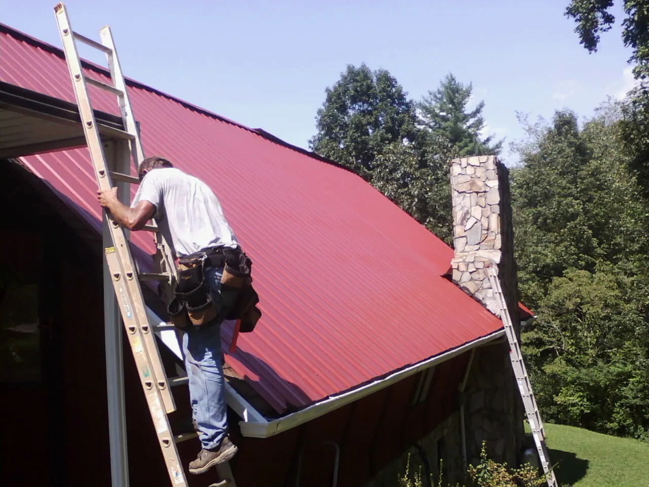 Roofer on a ladder beside a red metal roof and stone chimney — Soffit & Fascia Repair by Roofing Brothers in Rebecca